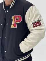 Campera estilo varsity con cuerpo negro y mangas blancas. Presenta parches bordados en el pecho y las mangas, cierre frontal con botones a presión y puños, cuello y ruedo acanalados con rayas blancas y negras.