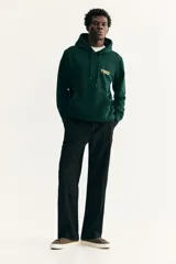 Canguro verde oscuro de peso medio con interior cepillado suave. Tiene bolsillo canguro, capucha de doble capa con cordón de ajuste y frente cruzado. Puños y bajo acanalados. Corte estándar. Estampado frontal con la palabra "VIBES" en amarillo.