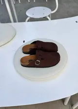 Zuecos de gamuza color marrón chocolate, con diseño cerrado en la parte delantera y tira ajustable con hebilla metálica en el empeine. Presentan plantilla anatómica de corcho y suela de goma.
