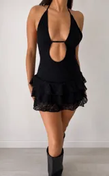 Vestido corto de encaje negro con diseño halter, escote profundo y abertura frontal. Presenta una falda con volados superpuestos y terminación en encaje transparente.