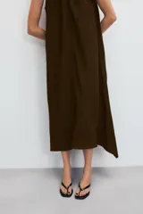 Vestido midi de color marrón con acabado satinado, cuello redondo con detalle fruncido y diseño sin mangas. Presenta un corte fluido con bajo asimétrico y bolsillos laterales ocultos.