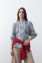 Camisa de rayas verticales con cuello mao, detalle de volados en el escote, bordados florales y recortes con calados. Posee mangas largas abullonadas con puños ajustados y cierre frontal con botones.