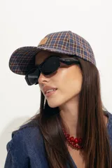 Gorra tipo baseball con estampado de cuadros escoceses en tonos azul, naranja y beige. Presenta un parche de cuero sintético en el frente y cierre ajustable en la parte posterior.