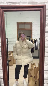 Campera puffer de corte oversize y diseño acolchado, en color beige. Presenta cuello alto, cierre frontal con solapa y botones a presión, y bolsillos laterales.