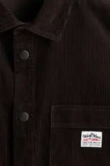 Camisa oversized de pana de algodón color marrón oscuro, con cuello inglés, cierre de botones a presión, dos bolsillos de parche en el pecho (uno con aplique), mangas largas con puños abotonados y bajo redondeado.