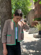 Chaqueta corta tejida a rayas horizontales marrones y blancas, con cuello redondo y ribete verde. Tiene bolsillos frontales y mangas 3/4.