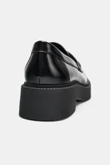 Mocasín de cuero negro con suela gruesa tipo plataforma, detalle de antifaz con aplique metálico dorado y costuras reforzadas en la puntera.
