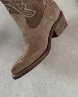 Bota corta de gamuza color marrón topo, estilo western, con bordados decorativos en hilo blanco en caña y empeine. Posee taco cuadrado de madera y punta ligeramente afilada.