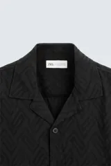 Camisa de corte relajado confeccionada en tejido fluido con mezcla de viscosa, color negro. Presenta cuello solapa, manga corta, cierre frontal con botones y un bordado a contraste en el pecho.