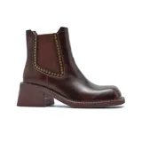 Bota tipo chelsea de cuero color marrón oscuro con acabado desgastado. Presenta paneles elásticos laterales decorados con tachas metálicas doradas, puntera cuadrada y taco bloque de altura media con textura amaderada.