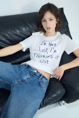 Camiseta de algodón de corte regular, con cuello redondo y manga corta. Presenta un estampado frontal con texto en color azul oscuro sobre fondo blanco.