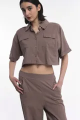 Camisa crop color chocolate, de corte holgado, con cuello clásico, cierre frontal de botones y dos bolsillos frontales con solapa. Presenta mangas cortas con dobladillo.