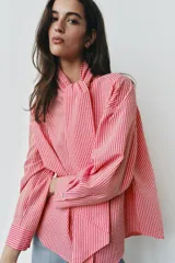 Camisa de rayas rosadas y blancas, con cuello subido decorado con un lazo integrado, manga larga con puños abotonados y cierre frontal oculto.