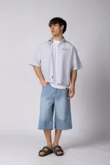 Camisa de manga corta de popelina rayada, con calce boxy y bordado sutil en el pecho. Presenta cierre frontal con botones y diseño abierto.