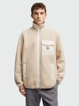 Buzo de polar tipo teddy en color beige, con cierre frontal completo, cuello alto y bolsillo en el pecho con solapa y logo de la marca. Presenta detalles en tela técnica al tono en los bordes de los bolsillos, cierre y cuello.