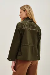 Campera estilo militar o safari color verde oliva, confeccionada en algodón. Presenta cuello solapa y bolsillos frontales con tapa. Destaca por sus apliques de pedrería o bordados en tonos marrones y dorados en el cuello y sobre los bolsillos.