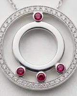 Collar de acero con cadena fina que presenta pequeños apliques de circonias a lo largo de su extensión. El dije central tiene un diseño circular doble, con el borde exterior pavé de circonias y tres pequeñas piedras rojas incrustadas en la parte inferior del círculo interior.