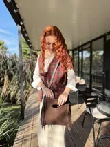 Cartera bandolera de cuero color chocolate, con forro interno de tela y flecos en la parte inferior. Tiene asa larga regulable y cierre superior con botón. Fabricada en Uruguay.