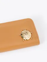 Billetera compacta de cuero vacuno color bordó, con cierre perimetral, correa de muñeca y aplique metálico dorado en forma de mariquita en el frente.