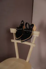 Zapatos tipo zueco de gamuza negra con punta fina y tira sobre el empeine decorada con tachas metálicas plateadas. Posee taco cuadrado de madera y suela con plataforma.