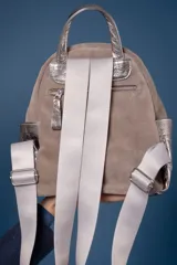 Mochila de cuero vacuno color tostado, con detalles en cuero metalizado plateado en los laterales y manija superior. Presenta un bolsillo frontal con cierre metálico, correas de cinta regulables y placa metálica con logo de la marca en la parte inferior.
