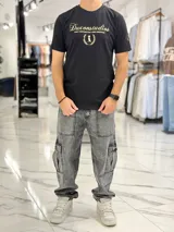 Remera color beige con cuello redondo y mangas cortas. Estampado frontal con el texto "Duxonstudios" en tipografía cursiva, acompañado de un logotipo circular con hojas de laurel y una figura central en tono verde oscuro.