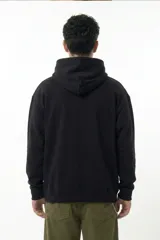Hoodie negro de corte relajado con capucha y cordones ajustables. Presenta un estampado gráfico central en el pecho con forma ovalada y bordes irregulares en tonos verde y rojo.