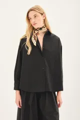 Camisa de popelina de algodón en color negro, con corte amplio, cuello clásico y cierre frontal con botones. Presenta un diseño versátil con botones en los laterales que permiten un ajuste cruzado.