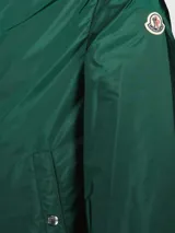 Campera cortavientos Moncler modelo Grimpeurs, color verde oscuro, con capucha ajustable, cierre frontal y bolsillos laterales con botones a presión. Presenta detalles de rayas tricolores en el interior de la capucha y puños elásticos.