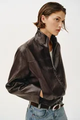 Chaqueta estilo bomber corta de efecto piel en color marrón oscuro, con cuello subido, cierre frontal con cremallera y cintura elástica fruncida.