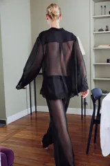 Blusa semitransparente negra de cuello solapa y manga larga, con cierre frontal de botones y detalles de bordados tonales en el ruedo y los puños.