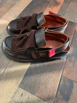 Mocasines de cuero color marrón oscuro, con diseño de flecos y tira con hebilla en el empeine, y suela plana con plataforma baja.