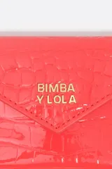 Monedero pequeño de estructura rígida, color negro con acabado brillante (efecto charol o vinilo). Presenta un cierre de solapa con botón a presión y correa de mano/muñeca. El logo metálico dorado de BIMBA Y LOLA está centrado en la solapa.