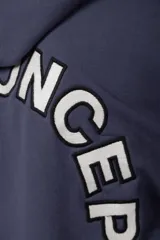 Buzo canguro oversize de frisa de algodón premium en color azul grisáceo. Presenta un diseño con recortes frontales, bolsillo tipo canguro y un pequeño logo circular estampado en el pecho. Cuenta con puños y ruedo acanalados.