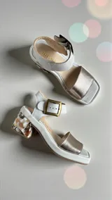 Sandalias de cuero color blanco y dorado, con taco ancho intervenido con arte pintado a mano, tira frontal metalizada, tira al tobillo con hebilla metálica y plataforma baja.