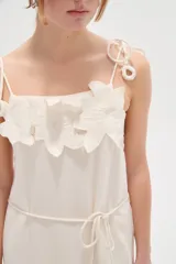 Vestido largo de algodón color blanco, con tirantes finos ajustables con lazos en los hombros, escote recto decorado con apliques florales tridimensionales, cordón ajustable en la cintura y falda larga con volados horizontales.
