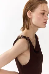 Vestido midi marrón sin mangas, de punto suave, con escote drapeado y cuentas decorativas irregulares en el tirante derecho. Presenta frunces en el lateral con efecto drapeado.