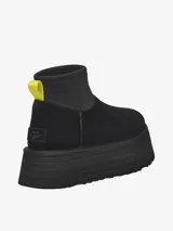 Bota corta tipo botineta de la marca UGG, color negro, con diseño de caña baja y plataforma elevada. Presenta una tira de agarre en color amarillo neón en la parte posterior y una suela de goma gruesa con textura acanalada.