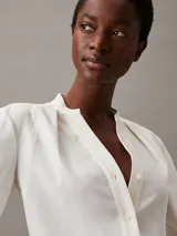 Blusa de manga larga color blanco roto, confeccionada en tejido Georgette de poliéster reciclado. Presenta un escote sin cuello con abertura en V y detalles plisados en los hombros y la espalda.