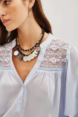 Camisa de algodón color celeste, con cuello mao, cierre frontal parcial con botones y detalles de encaje en los hombros y canesú. Presenta mangas largas abullonadas con puños elásticos.