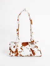 Cartera tipo baguette confeccionada en cuero con pelo con estampado animal print de vaca en tonos blanco y marrón, con doble asa larga para el hombro.