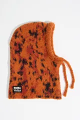 Balaclava de punto con diseño envolvente, confeccionada en tejido texturizado con estampado animal print tipo lince en tonos naranja, rojo y negro. Presenta cordones ajustables en el cuello y etiqueta con logo de la marca en la parte inferior.