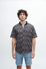Camisa de manga corta con cuello camisero y botones frontales. Presenta un estampado tipo paisley o mandala en tonos morados, verdes y marrones sobre un fondo oscuro.