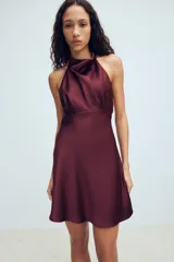 Vestido corto de tejido satinado color bordó, con cuello halter, espalda descubierta y detalle de frunces en la cintura.