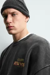 Sudadera de corte relajado, color negro con efecto lavado, cuello redondo y manga larga. Presenta un pequeño estampado frontal con texto en tonos naranja y amarillo.