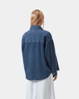 Camisa de denim azul de corte holgado, con cuello clásico, cierre frontal de botones metálicos y bolsillo parche en el pecho con etiqueta cuadrada de la marca.