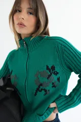 Cardigan de punto color marrón con cierre frontal, cuello alto y bolsillos aplicados. Presenta diseño tejido localizado con detalles en violeta y negro en el pecho.