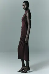 Vestido midi de color negro, con cuello subido y diseño sin mangas. Presenta un detalle de drapeado lateral en la parte delantera y cierre de cremallera oculta en la espalda.