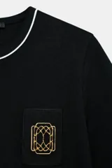 Camiseta negra de algodón, corte regular, con cuello redondo ribeteado en blanco y un bolsillo frontal con un broche metálico decorativo dorado.