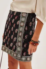 Falda corta negra estilo wrap con estampado paisley en tonos rojo y blanco. Presenta un borde decorativo con motivos circulares en tonos plateados y beige.